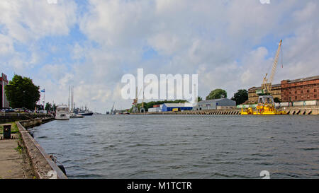 Harbor with yellow crane on a barge in front of a quay with train freight cars and abandoned industrial buildings on one side and leisure sailing boat Stock Photo