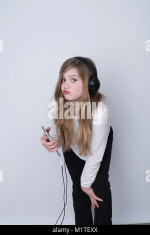 Girl with pouty look listening to music on smartphone with over-the-ear headphones on white background Stock Photo