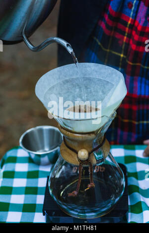 Barista pours boiling water fron specialized kettle on pour over coffee in artisan glass brewer while outdoors Stock Photo