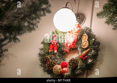 Beautiful Christmas decorations hanging on doors outside the house. Green needle crown with pine cones, angels, dry fruits, elf and red ribbons. Stock Photo