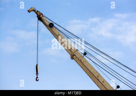 Shot of yellow boom with big hook, an old, rusty port crane preparing to lift cargo in ship on clear blue sky background. Equipment for unloading ship Stock Photo
