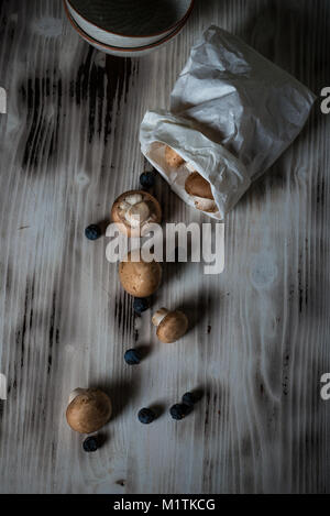 Vertical photo with top view on spilled brown edible mushrooms from paper bag. The champignons are on vintage wooden board with white worn color and   Stock Photo