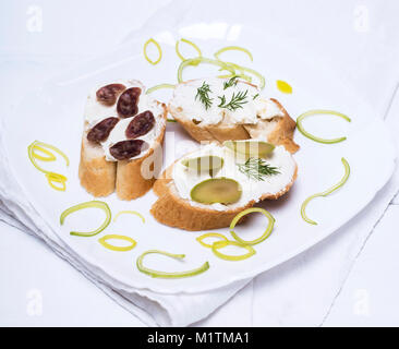 sandwiches with creamy white cheese, sausage, olives and dill on a white plate, wooden white table Stock Photo