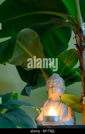 Sitting Buddha surrounded by banana trees hold the burning candle in hands Stock Photo