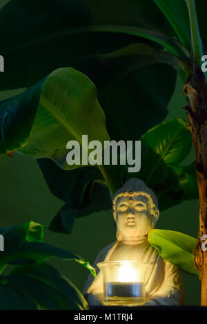 Sitting Buddha surrounded by banana trees hold the burning candle in hands Stock Photo