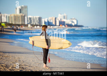 MIAMI - JANUARY 4, 2018: A young female surfer in a wet suit stands on the shore of South Beach with her surfboard looking out at the winter swells. Stock Photo