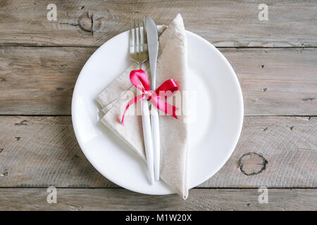 Romantic table setting with white plate, modern cutlery and red ribbon on wooden background, top view, copy space. Romantic dinner, dating, wedding co Stock Photo