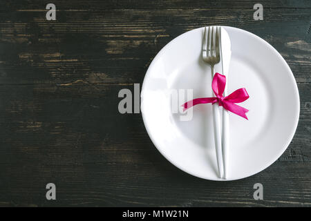 Romantic table setting with white plate, modern cutlery and red ribbon on dark black wooden background, top view, copy space. Romantic dinner, dating, Stock Photo