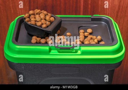 Small pieces of dry complete dog food / kibble in a measuring scoop and around the lid of a green and black plastic storage bin / container. UK. Stock Photo
