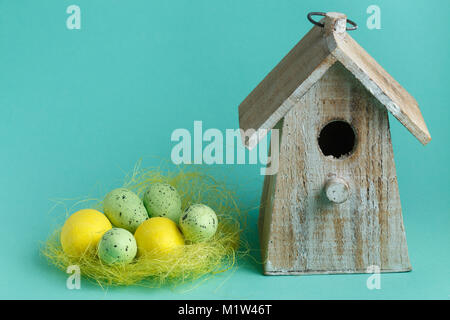 color Easter eggs in nest on turquoise background with wooden vintage birdhouse. Stock Photo