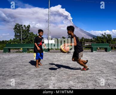 Daraga, Albay, Philippines. 23rd Jan, 2018. Children play basketball on a recreation yard in Daraga while the Mayon volcano erupts Tuesday. The Mayon volcano continued to erupt Tuesday, although it was not as active as it was Monday. There were ash falls in communities near the volcano. This is the most active the volcano has been since 2009. Schools in the vicinity of the volcano have been closed and people living in areas affected by ash falls are encouraged to stay indoors, wear a mask and not participate in strenuous activities. Credit: Jack Kurtz/zReportage.com/ZUMA Wire/Alamy Live News Stock Photo