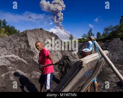 Daraga, Albay, Philippines. 23rd Jan, 2018. Sand and gravel miners work in a riverbed of volcanic soil while the Mayon volcano erupts Tuesday. The Mayon volcano continued to erupt Tuesday, although it was not as active as it was Monday. There were ash falls in communities near the volcano. This is the most active the volcano has been since 2009. Schools in the vicinity of the volcano have been closed and people living in areas affected by ash falls are encouraged to stay indoors, wear a mask and not participate in strenuous activities. (Credit Image: © Jack Kurtz/zReportage.com via ZUMA Wi Stock Photo