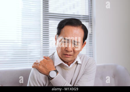 Elderly man suffering from shoulder pain sitting on a sofa in the living room Stock Photo