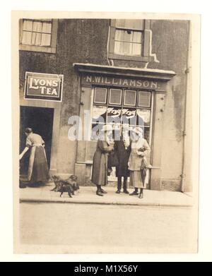 Early 1920s photograph of two women chatting with a man outside a grocer's shop, there is an advertising sign for Fry's Cocoa in the shop window and a Lyon's Tea advert above the shop doorway. An older woman still wears a long skirt in Edwardian style, whilst the two women chatting have adopted a higher hemline, U.K. Stock Photo