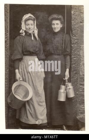 Original charming Edwardian era postcard portrait photograph of milk maid holding a milk pail and wearing a bonnet on the doorstep of a house with a lady customer, borough of Rosendale, Rawtenstall, Ramsbottom area, U.K. circa 1906 Stock Photo
