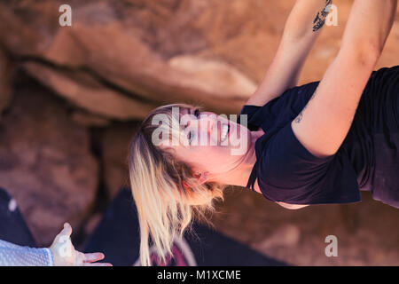 Thin blonde caucasian woman with arm tattoos hangs upside down while she rock climbs on boulders in the desert of California Stock Photo
