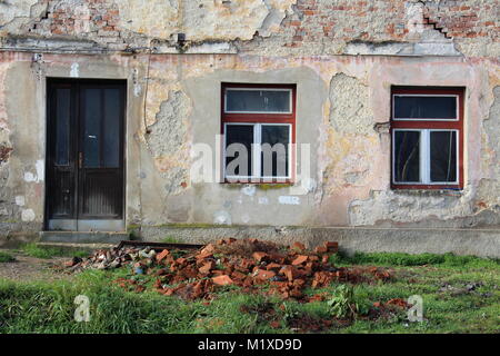 Abandoned old ruined house closeup of dilapidated front doors with missing door knob, two windows, partially fallen facade and red bricks in front on Stock Photo