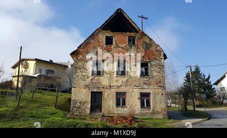Abandoned old ruined house with broken windows, fallen facade and bricks from walls, next to paved road and with modern suburban house in background Stock Photo