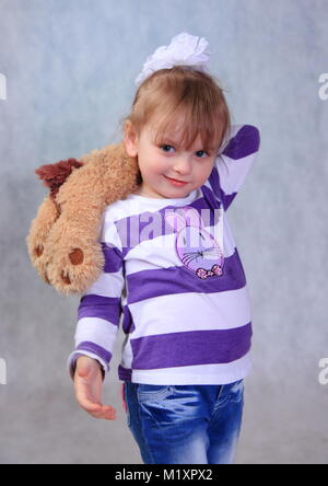On a gray background stands a little girl with a dog in his hands. She is dressed in a jacket and jeans. Stock Photo