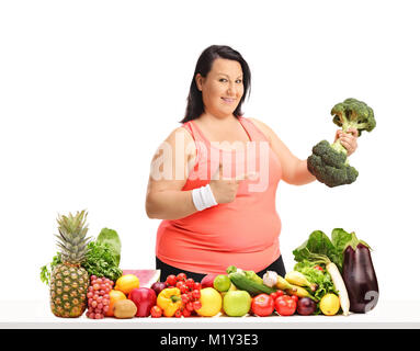 Overweight woman with a broccoli dumbbell pointing behind a table with fruit and vegetables isolated on white background Stock Photo