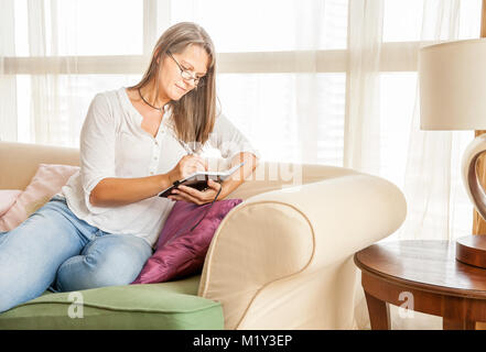 Mature woman is making notes in her diary sitting on a couch in her apartment Stock Photo
