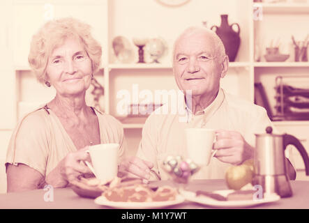 Smiling elderly spouses enjoying time at home, drinking tea with sweets Stock Photo