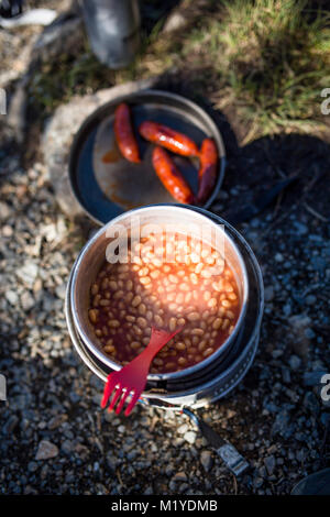 Baked beans in a saucepan and sausages in a frying pan on a camping kitchen on the ground. Partly sun and partly shadow. Stock Photo