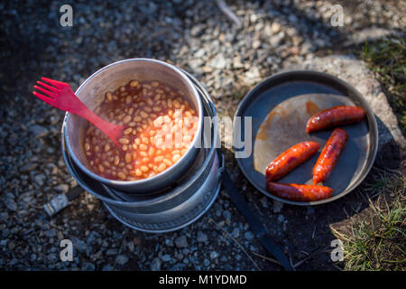 Baked beans in a saucepan and sausages in a frying pan on a camping kitchen on the ground. Partly sun and partly shadow. Stock Photo