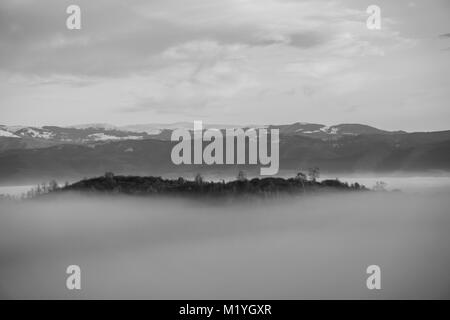 Black and white shot of small hill piercing the thick fog that engulfed the valley below Stock Photo