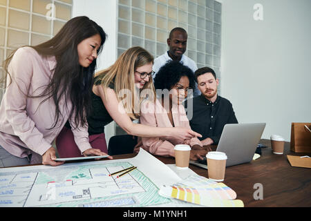 Diverse group of designers smiling and talking while working together over a laptop at a desk in a modern office Stock Photo
