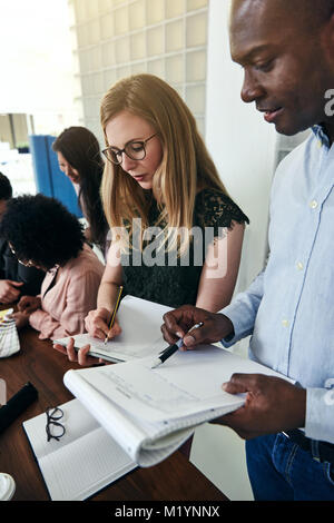 Two focused colleagues comparing notes while standing together in a modern office with coworkers working in the background Stock Photo