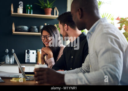 Three diverse coworkers smiling and talking together while sitting at a boardroom table during a meeting in a modern office Stock Photo