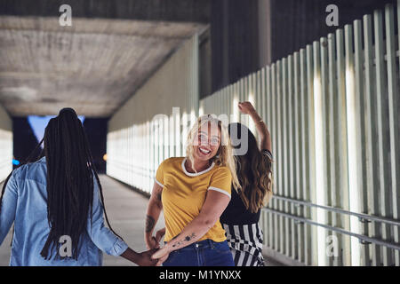 Laughing young woman walking hand in hand with two girlfriends down a walkway in the city at night Stock Photo