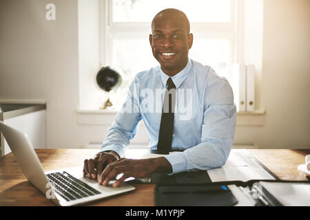 Successful young African businessman sitting at his desk in an office smiling and working on a laptop Stock Photo