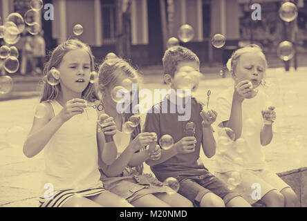 Portrait of positive girls and boy sitting at yard and blowing soap bubbles Stock Photo