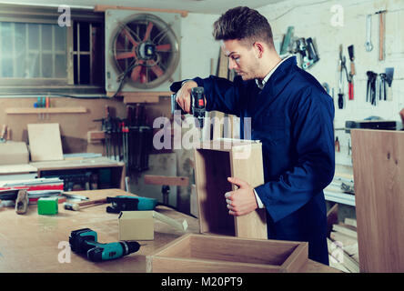 Young positive guy using drilling machine to make drawer at workshop Stock Photo