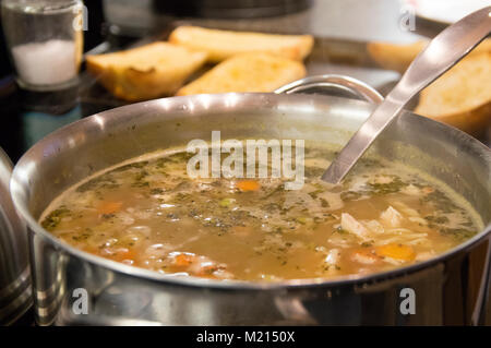 Homemade chicken noodle soup with vegetables in a large stainless steel pot with toasted bun slices in the background Stock Photo