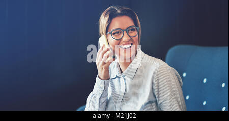 Young businesswoman laughing while sitting on a sofa at home talking on her cellphone Stock Photo