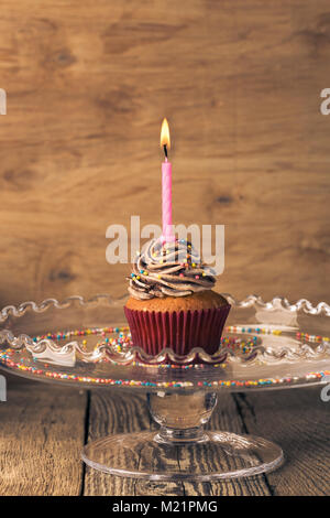 Delicious birthday cupcake with one candle served on glass cakestand on festive wooden table, retro toned Stock Photo