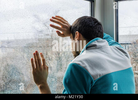 Depressed young man upset with bad news looking through rainy window glass Stock Photo