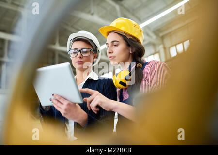 Portrait of two female engineers wearing hardhats standing in spacious workshop of modern plant using digital tablet Stock Photo