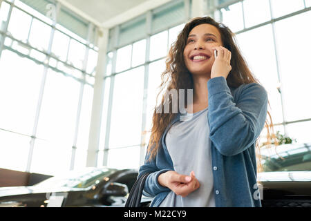 Low angle view of attractive mixed race woman looking away with charming smile and talking to her friend on smartphone while choosing new car in showr Stock Photo