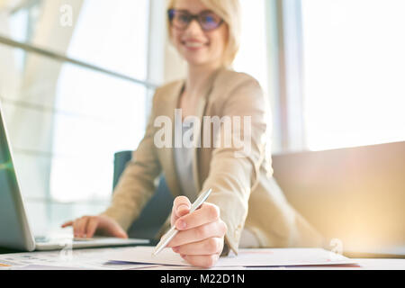 Cheerful young manager wearing eyeglasses looking at camera with toothy smile while distracted from work in spacious office with panoramic windows, fo Stock Photo