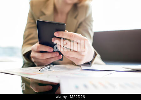 Close-up shot of unrecognizable white collar worker sitting at coffee table of office lobby and browsing Internet on smartphone, blurred background Stock Photo