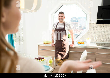 Portrait of happy young couple talking in modern kitchen lit by sunlight, focus on smiling young man wearing apron looking at his girlfriend across ro Stock Photo