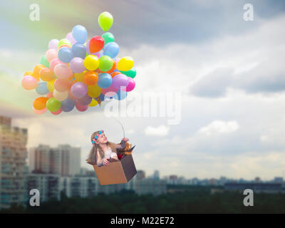 Happy girl flying in a cardboard box on the balloons. Flying high above the city. The concept of children's dreams, journey, freedom Stock Photo