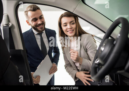 Portrait of cheerful young woman looking inside car while choosing luxury car in dealership showroom standing next to handsome salesman Stock Photo