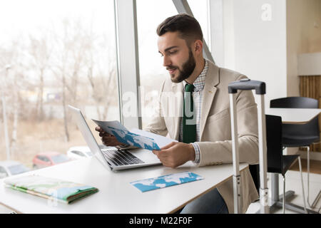 Portrait of handsome bearded man checking plane tickets sitting at table against window in airport, copy space Stock Photo