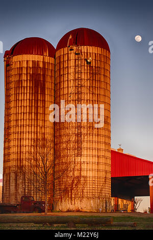 Vertial photo of two silos colored by the sunset with an old vehicle and a red roof with a bright blue sky and full moon in the background Stock Photo