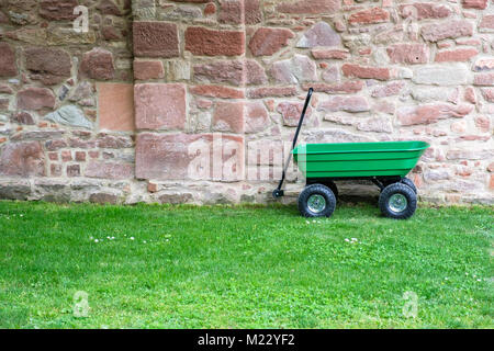 A green garden pushcart, wheelbarrow on the green grass field with old brick wall in the background. Stock Photo